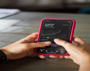 Close-up of hands holding a pink smartphone displaying an investing app with a green line chart and $891.26 label.