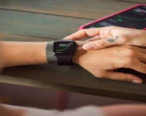 Wrist with a black smartwatch showing a green stock chart and price on a wooden table, with a pink-edged phone in the background.