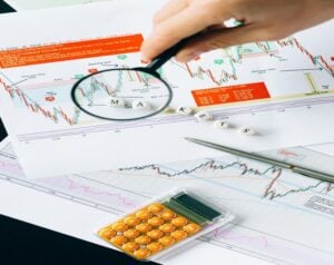 Analyst uses a magnifying glass to examine a stock chart with candlesticks; a calculator and pen nearby.