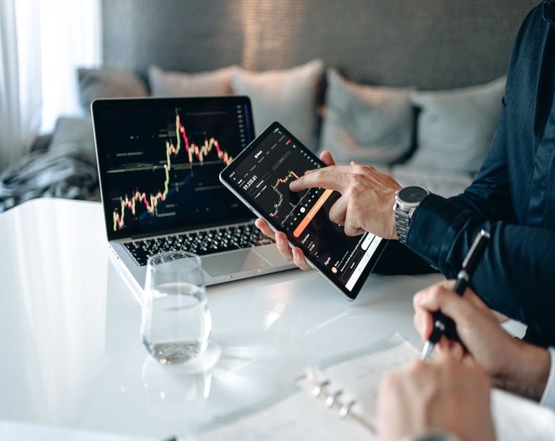Person monitors stock charts on a tablet while a laptop displays a stock chart on a bright desk at home.