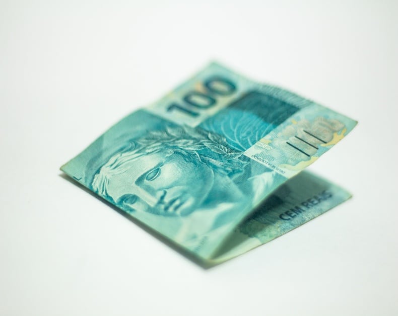 Blue-green 100 Brazilian real banknote partially folded on a white surface, showing the portrait and security features.