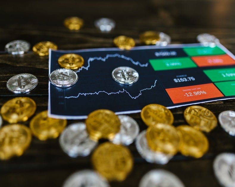 Close-up of a crypto trading screen with a fluctuating chart, surrounded by gold and silver coins on a wooden table.