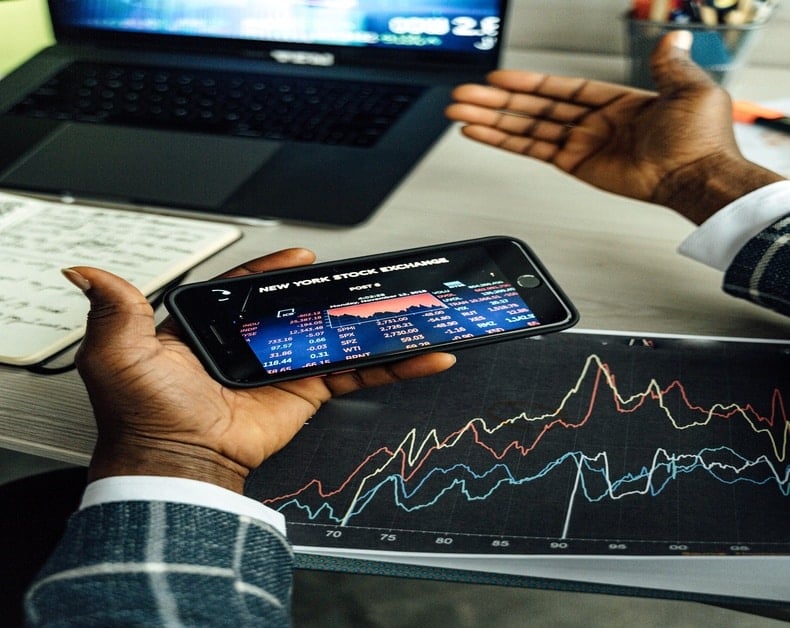 Hands hold a smartphone showing NYSE stock quotes; a colorful stock chart is on the desk with a laptop nearby.