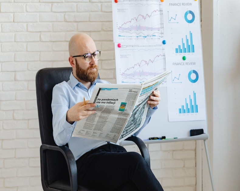 Bearded man with glasses sits in an office chair reading a newspaper, with financial charts on a whiteboard behind him.