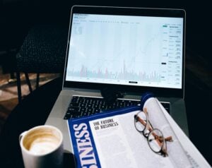 Laptop screen shows a rising stock chart with bars and metrics on a dark table, beside a business newspaper, glasses, and a coffee mug.