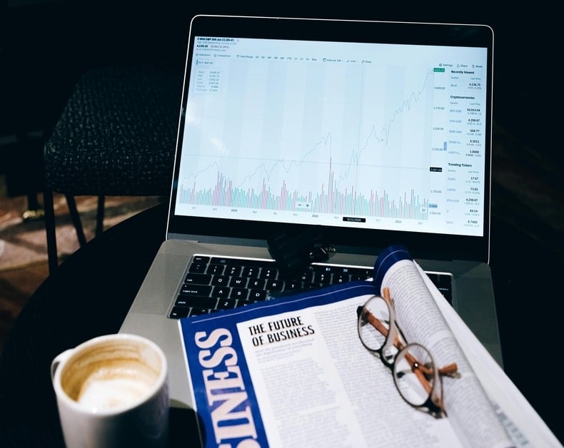 Laptop screen shows a rising stock chart with bars and metrics on a dark table, beside a business newspaper, glasses, and a coffee mug.