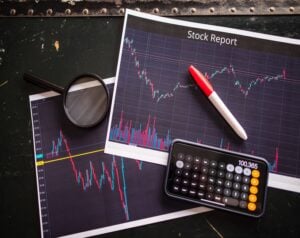 Stock report charts with a red pen, magnifying glass, and a calculator on a dark desk, conveying financial analysis visuals.