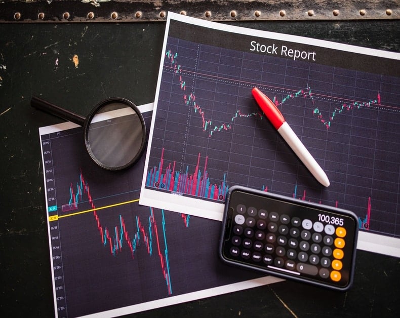 Stock report charts with a red pen, magnifying glass, and a calculator on a dark desk, conveying financial analysis visuals.