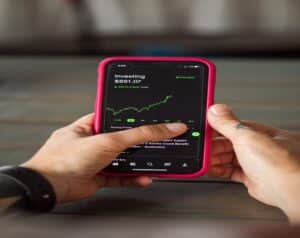 Person holding a phone in a pink case displaying an investing app with a green stock chart on screen.