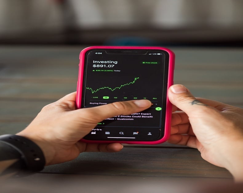 Person holding a phone in a pink case displaying an investing app with a green stock chart on screen.