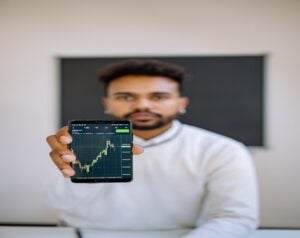 Man in a white shirt extends a smartphone showing a green candlestick stock chart, with a blurred background behind him.
