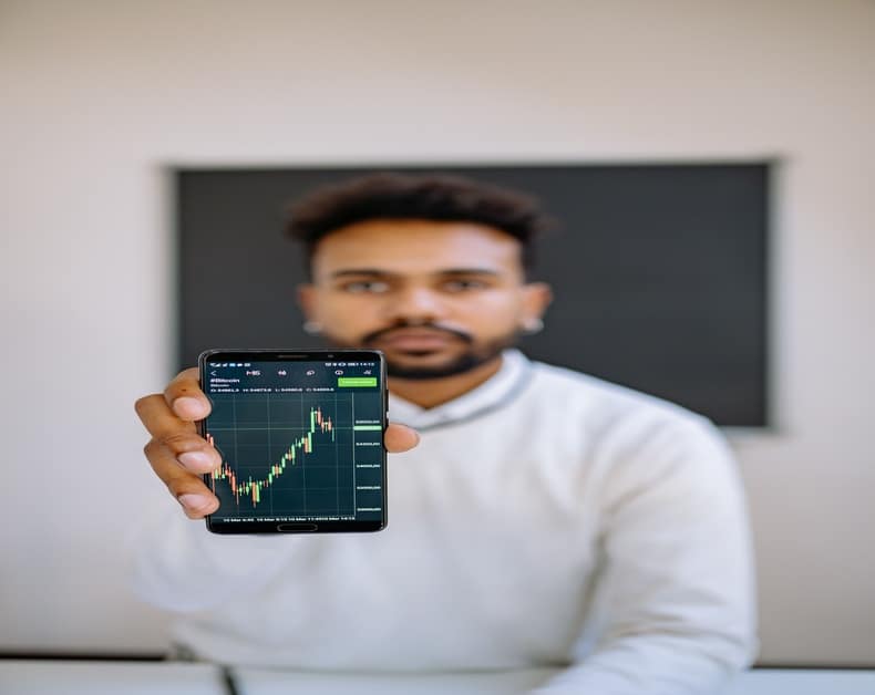 Man in a white shirt extends a smartphone showing a green candlestick stock chart, with a blurred background behind him.