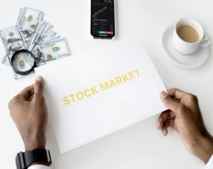 Hands holding a STOCK MARKET sheet with cash, a magnifying glass, a charted phone, and a coffee cup on a white desk.