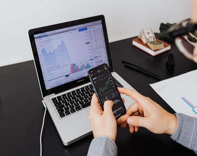 Person holding a smartphone displaying a stock chart while a laptop shows financial graphs on a desk