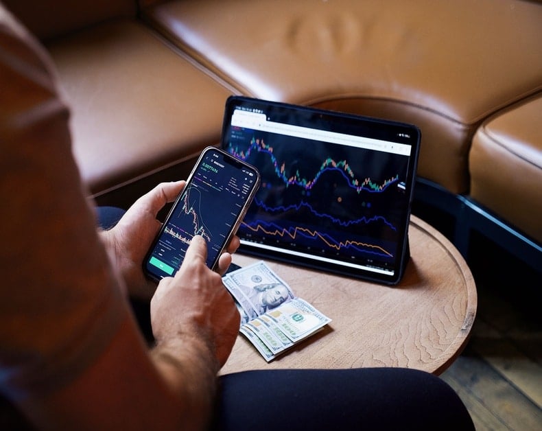 Person holds a smartphone displaying stock charts while a tablet shows market graphs on a round table with cash nearby.