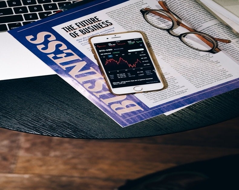 Phone showing a stock chart resting on a business newspaper with reading glasses nearby.