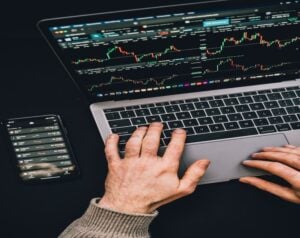 Person typing on a laptop displaying trading charts and a smartphone with market data beside it; financial monitoring setup.