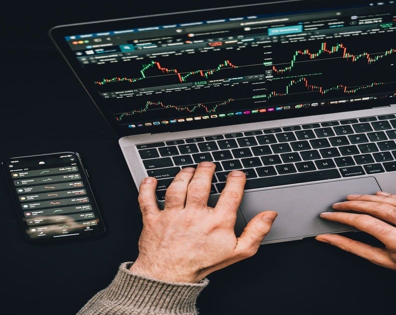Person typing on a laptop displaying trading charts and a smartphone with market data beside it; financial monitoring setup.