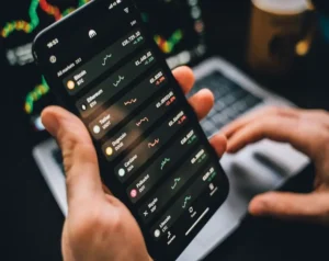 Person holding a smartphone in dark mode displaying a cryptocurrency price list with green and red trend lines.