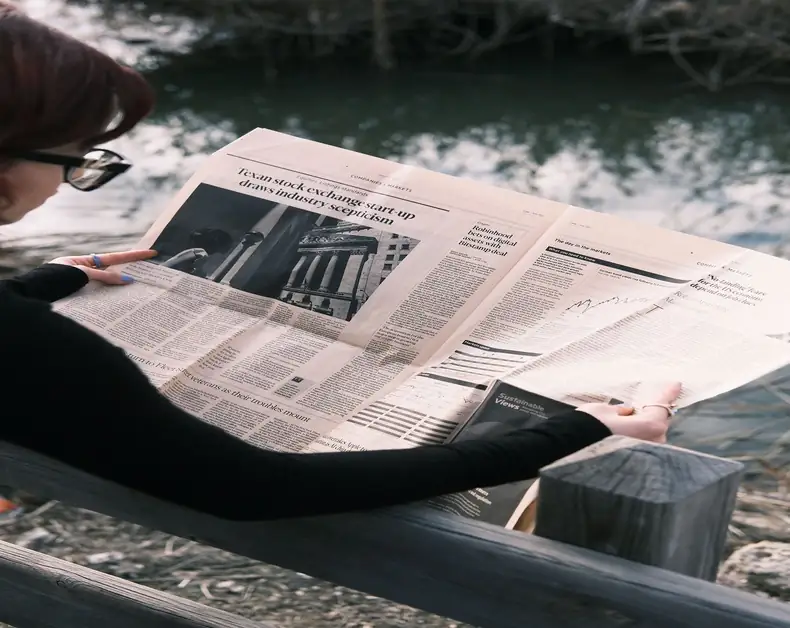Person wearing black sleeves and glasses reads a newspaper on a wooden bench by a lake.