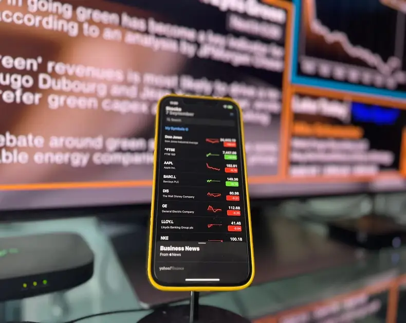 Smartphone with a stock market app showing red and green price changes on a dark screen, yellow case, in focus; blurred monitors with charts in the background.
