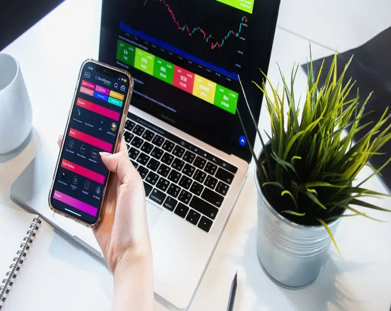 Person holds a smartphone displaying a colorful trading app next to a laptop showing a stock chart on a desk with a plant nearby.
