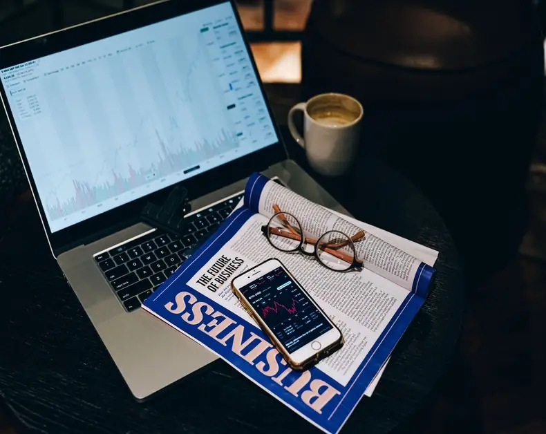 Laptop showing a financial chart on screen with a mug nearby, glasses on a business magazine and a smartphone displaying stock data.