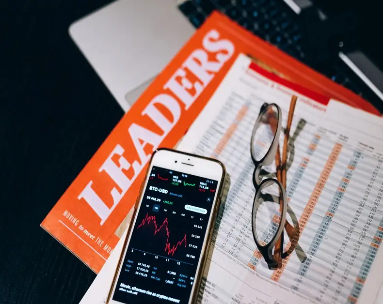 Phone shows a stock chart beside an orange Leaders magazine and reading glasses on a desk with a financial newspaper/printout nearby.