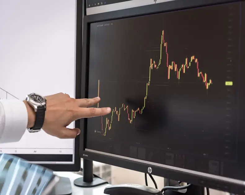 Person pointing at a candlestick stock chart on a computer monitor.